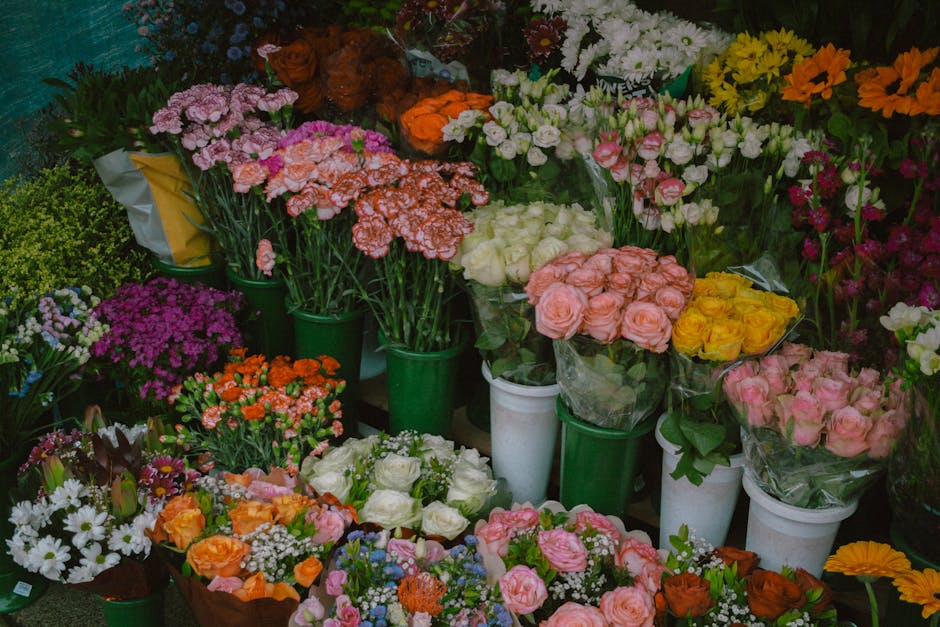 An array of vibrant fresh flowers arranged in white and green pots and bouquets, displayed on a table. The collection includes pink and peach roses, white roses, yellow chrysanthemums, pink blossoms, 