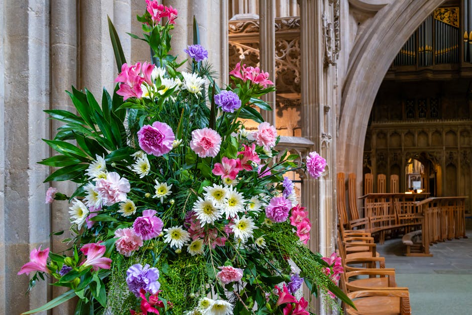 A large, vibrant floral arrangement positioned inside a historic church, featuring a lush mix of pink carnations, white daisies, purple limonium, and pink lilies, accented with green foliage. The bouq