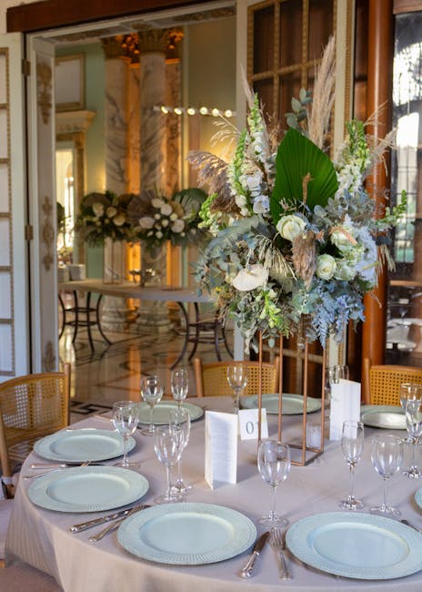 A formal dining table set with white tablecloths and elegant glassware, featuring a tall, lush floral arrangement as the centerpiece. The bouquet includes white roses, soft green foliage, large broad 